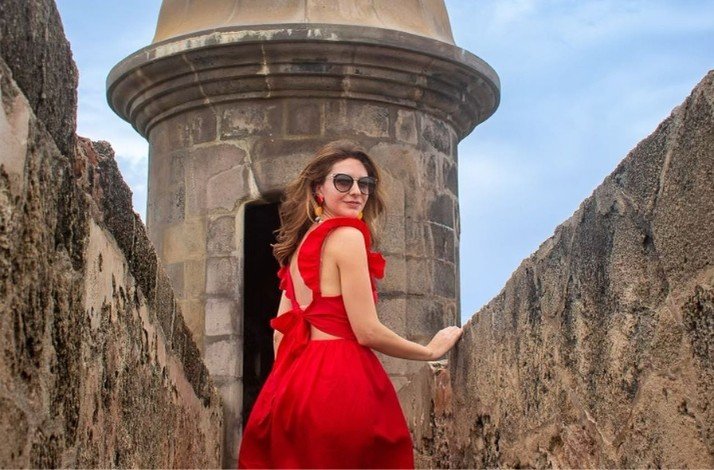 A woman in a red dress stands atop a stone fortress, posing for a picture against a backdrop of the Castillo San Felipe del Morro.
