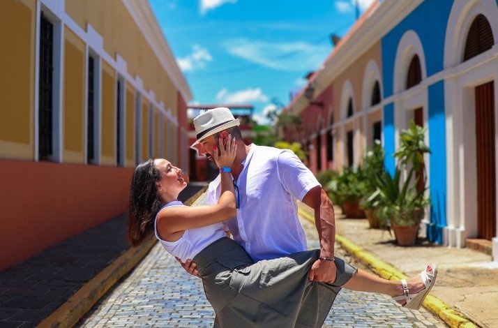 A man lifts a woman in a loving embrace, strolling down a colorful street of San Juan lined with pastel-colored buildings.