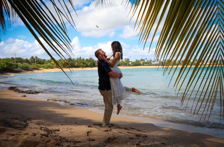 A couple stands on a beach, the man lifting the woman in a joyous embrace, bathed in the warmth of a sunny day in San Juan.