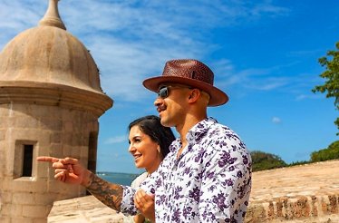 A couple stands at the Castillo San Felipe del Morro, pointing towards a distant vista under a clear blue sky.
