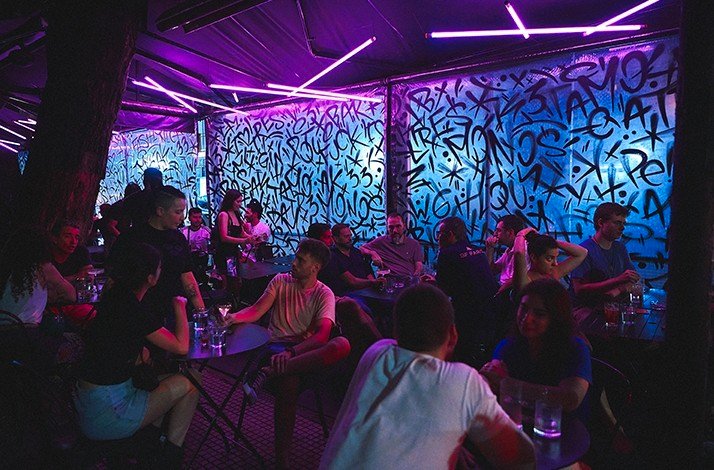 A vibrant TRES MONOS bar with neon purple lighting and graffiti-covered walls, filled with people socializing at tables.