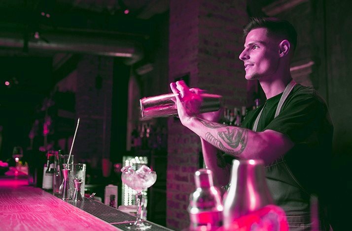 A bartender shaking a cocktail shaker behind a bar illuminated with pink lighting.