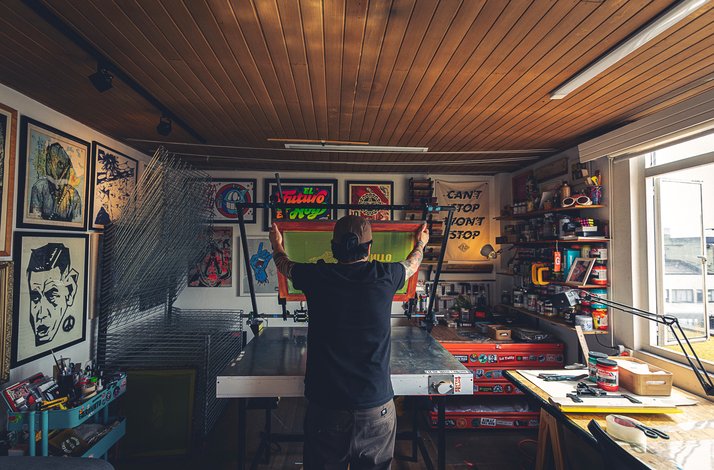 Designer lifting a screen-print frame in a studio surrounded by colorful prints and tools
