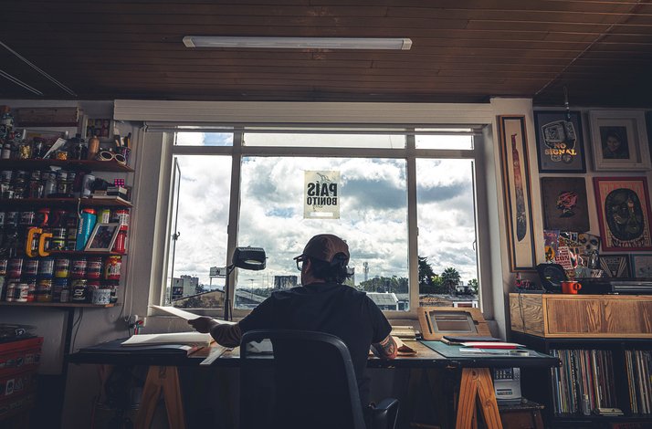 Artist working at a desk by a sunlit window with art tools and materials around
