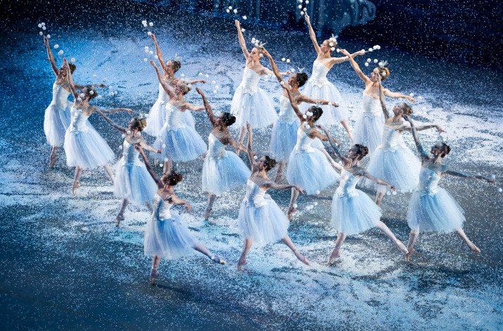 Ballet dancers dressed as snowflakes performing a synchronized routine in falling snow.