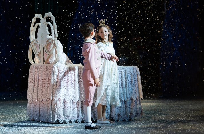 Young Clara and the Prince on stage in a snowy scene with a decorative bed.