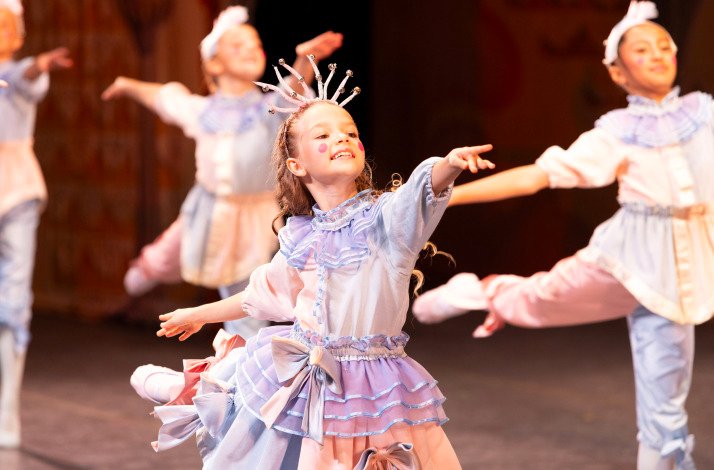 A smiling young Clara in pastel costume dances joyfully in a group performance.