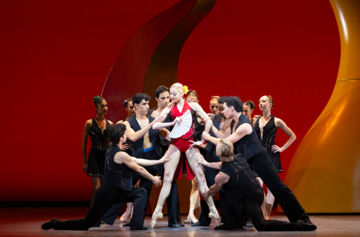 Group of dancers surround central figure in red, dynamic stage interaction.