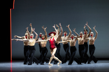 Ballet dancers in dramatic pose, center in red costume, arms raised on stage.