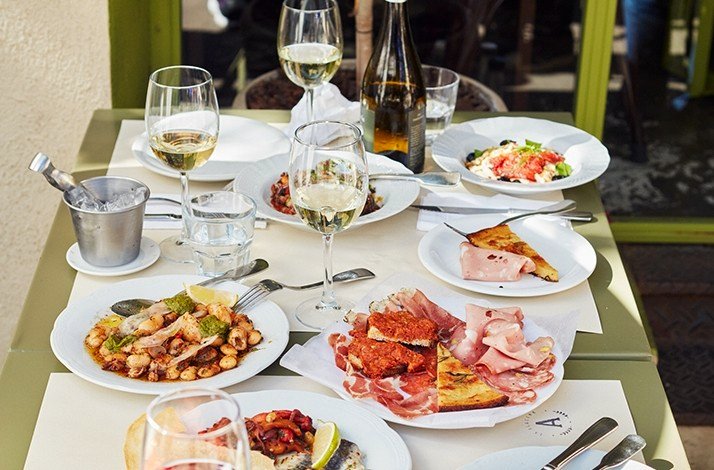 La Alacena Trattoria's outdoor table is set with various dishes of food, including pasta, salads, and meats, along with glasses of wine