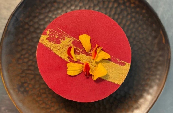 A vibrant dessert with a red and gold design, adorned with yellow flower petals on a textured black plate
