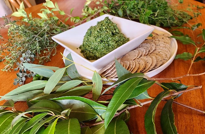 A bowl of green pesto is served with crackers on a wooden table surrounded by various types of leaves.