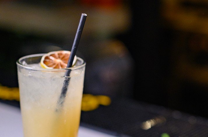 A view of a glass of amber-colored cocktail on a dark wooden surface, with a vintage radio and books in the background.