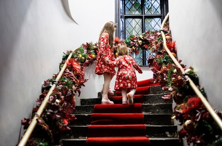 Two young girls in red Christmas-themed dresses are walking up a staircase decorated in red carpet and Christmas ornaments.