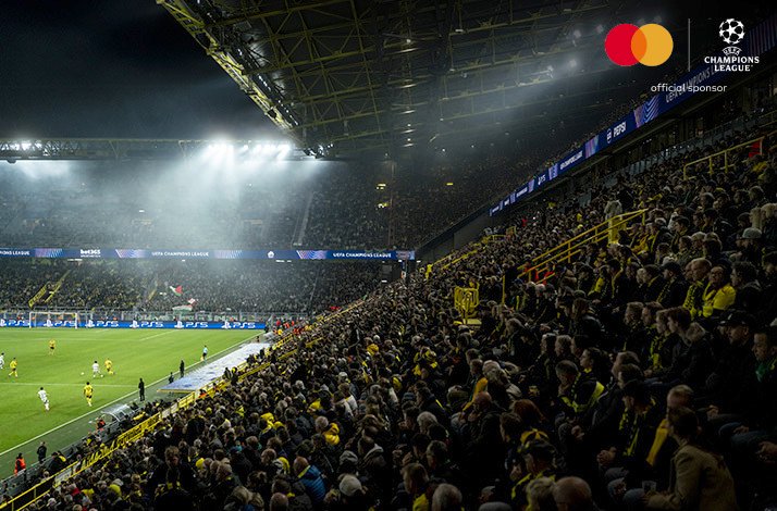 Packed stadium crowd cheering under floodlights during a UEFA Champions League night match, with Mastercard official sponsor logo in upper-right corner.