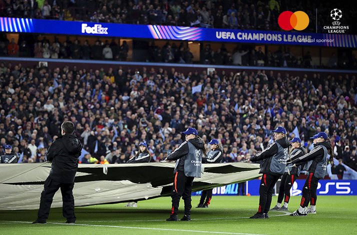 Ground staff unfolding a large banner on the pitch at a UEFA Champions League match before a packed stadium crowd., on upper right corner Mastercard logo + UEFA Champions League logo with text 'official sponsor'.