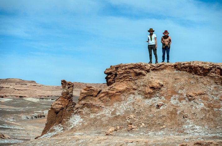 Two people with cowboy hats standing on a large rock formation in Salt Mountain Range, San Pedro de Atacama, Chile.