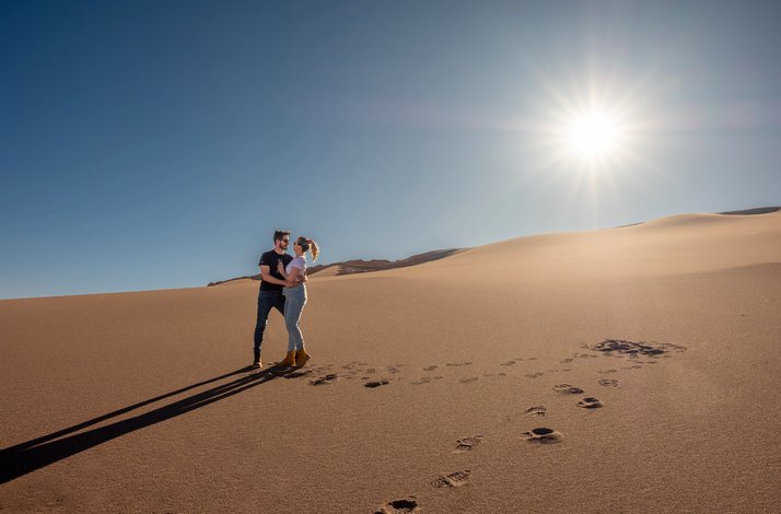 A couple holding each other in a sand valley while the sun shines behind them.