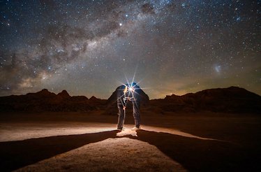 Two figures stand in Atacama Desert under the night sky filled with stars and the Milky Way, illuminated by a light source. 