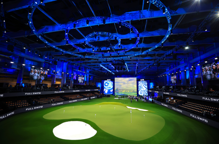 Modern indoor golf arena with putting greens, digital screens, and dramatic blue lighting.