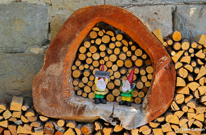 Two garden gnome figurines placed inside a hollow log, surrounded by stacked firewood