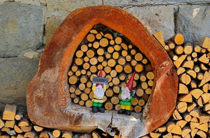 Two garden gnome figurines placed inside a hollow log, surrounded by stacked firewood