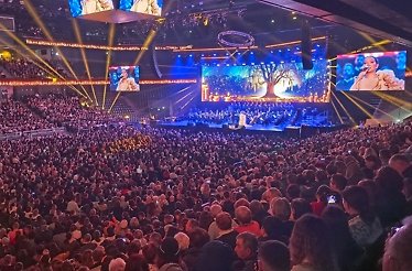 Packed auditorium with vibrant stage lights, a choir, and a tree-themed backdrop during a live performance