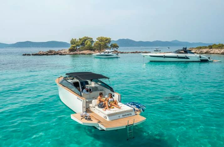 A couple relaxes on a deck of a white motorboat in a turquoise bay, surrounded by islands and other boats at Dubrovnik. 