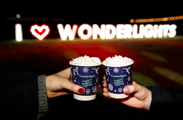 Two hands toasting with festive hot drinks in front of a glowing I love WONDERLIGHTS sign.