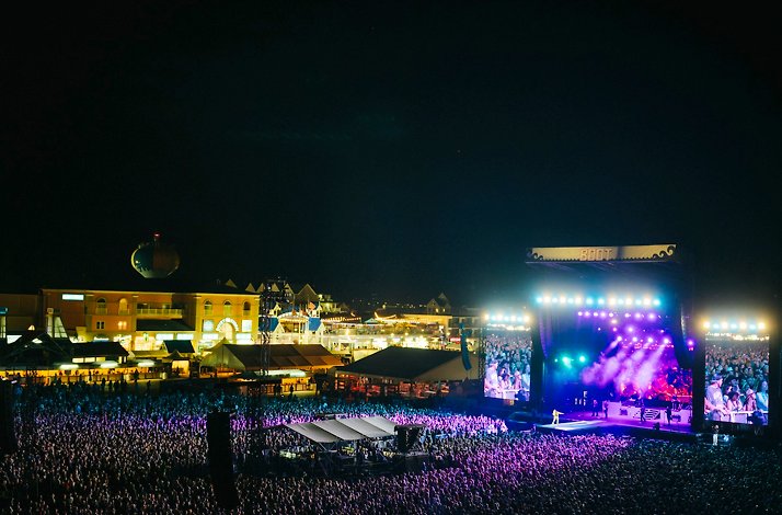 Large crowd gathers at an outdoor music festival by the waterfront, with a brightly lit stage and surrounding buildings at night.