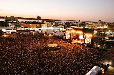 Large crowd gathers at an outdoor music festival by the waterfront, with a brightly lit stage at sunset.