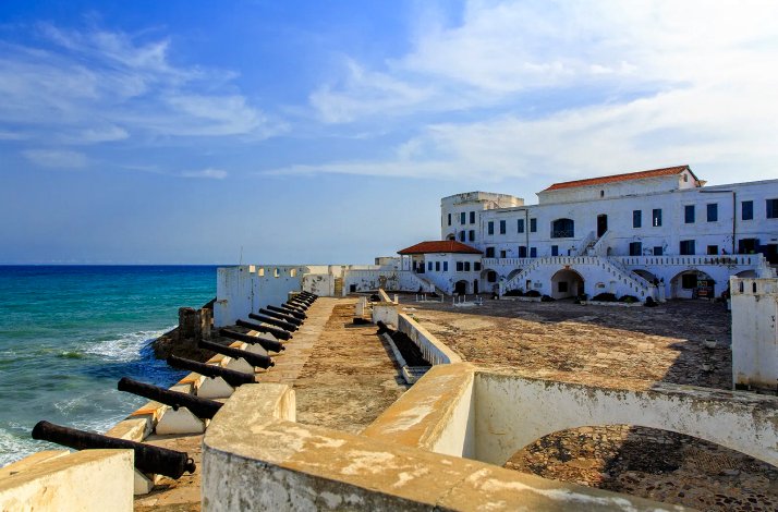 A historic Cape Coast Castle with cannons lined along its walls, adjacent to the ocean