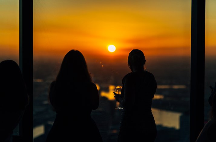 Two guests silhouetted against floor-to-ceiling windows at Eureka 89, overlooking Melbourne's Southbank skyline at sunset, one holding a wine glass.