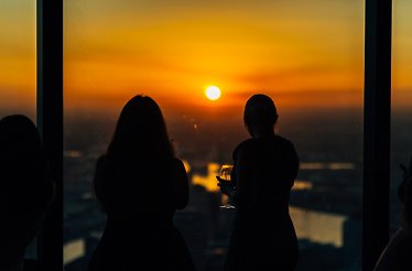 Two guests silhouetted against floor-to-ceiling windows at Eureka 89, overlooking Melbourne's Southbank skyline at sunset, one holding a wine glass.