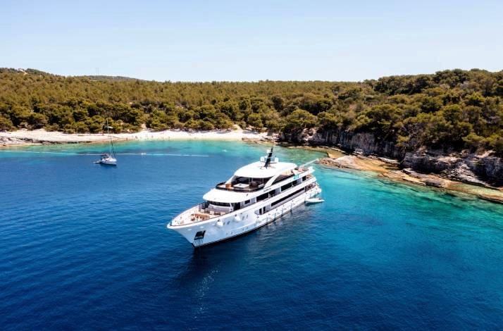 A luxurious white yacht is anchored in the blue waters of Croatian Adriatic, surrounded by lush green forested shores.