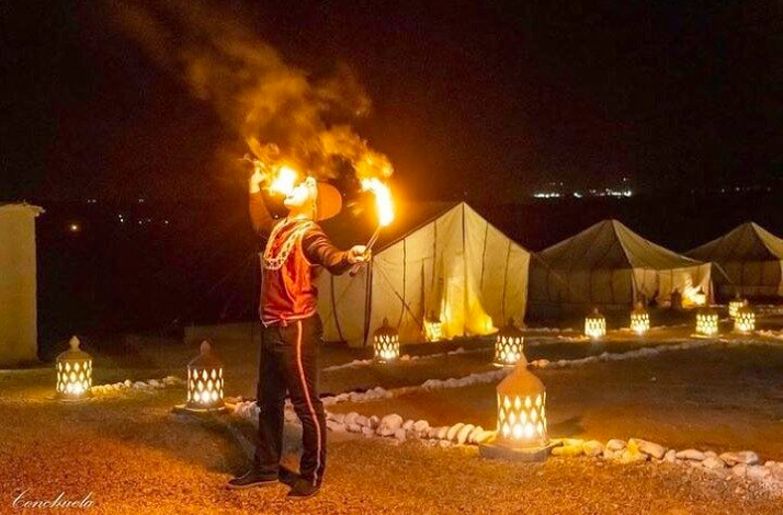 A nighttime scene at Agafay Desert captures a performer breathing fire amidst illuminated lanterns and tents.