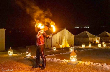 A nighttime scene at Agafay Desert captures a performer breathing fire amidst illuminated lanterns and tents.