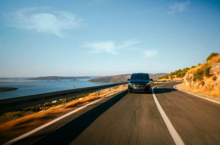 Dark premium transfer van travelling a sun-lit Croatian highway overlooking island-dotted Adriatic waters under clear skies. 