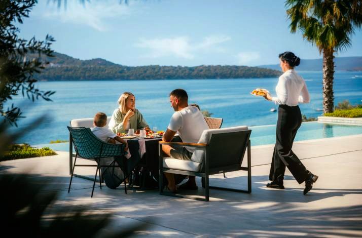 Server presenting a dish to a family dining on a sunlit terrace overlooking an Adriatic bay beside a private villa pool.