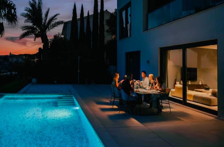 Friends gathered around an outdoor dinner table beside an illuminated villa pool at twilight in Croatia. 