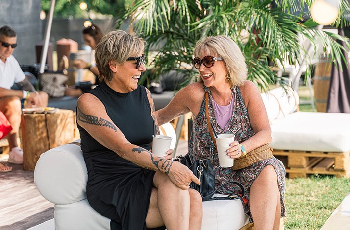 Two women relax on outdoor lounge chairs, enjoying drinks at the festival