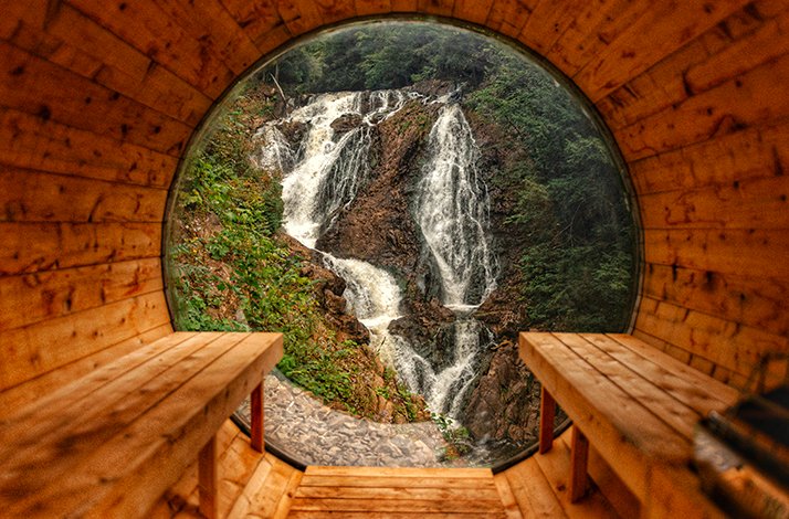 A view of a cascading waterfall framed by a wooden sauna's circular window, at Calumet Falls