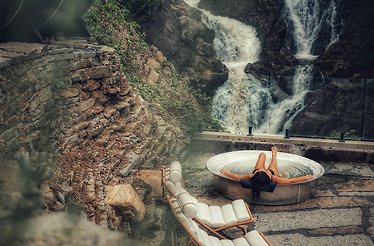 A woman relaxes in a circular outdoor tub with a waterfall cascading nearby, at Calumet Falls