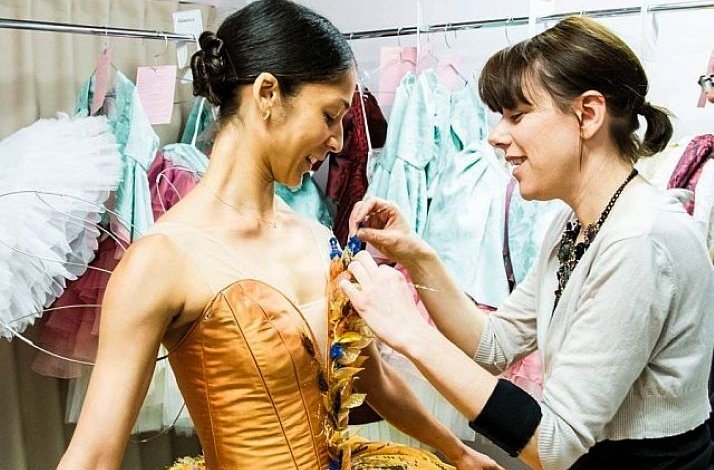 A costume designer adjusting a ballet dancer's ornate golden corset in a backstage dressing room.