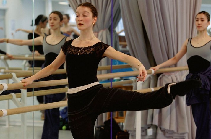 Dancers at the barre in a mirrored studio, practicing leg extensions.