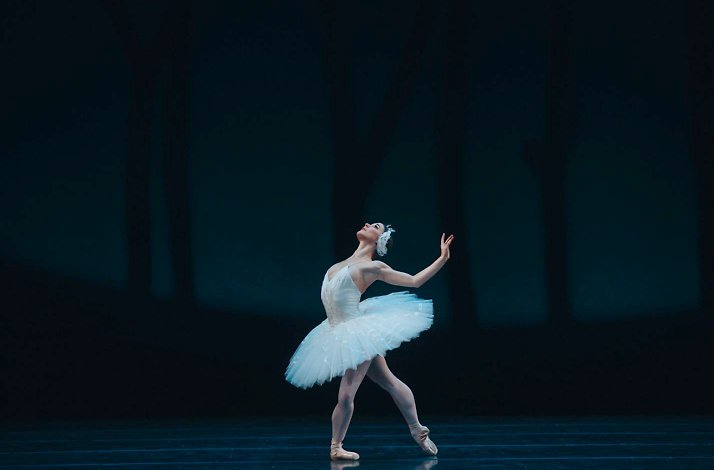 Ballerina in a white tutu performing a graceful pose on a dimly lit stage.