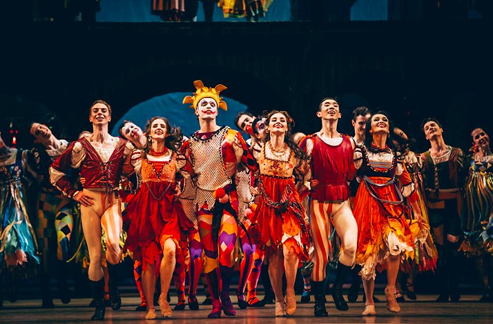 A troupe of dancers in vibrant Renaissance carnival costumes with a central jester in yellow from Romeo and Juliet.