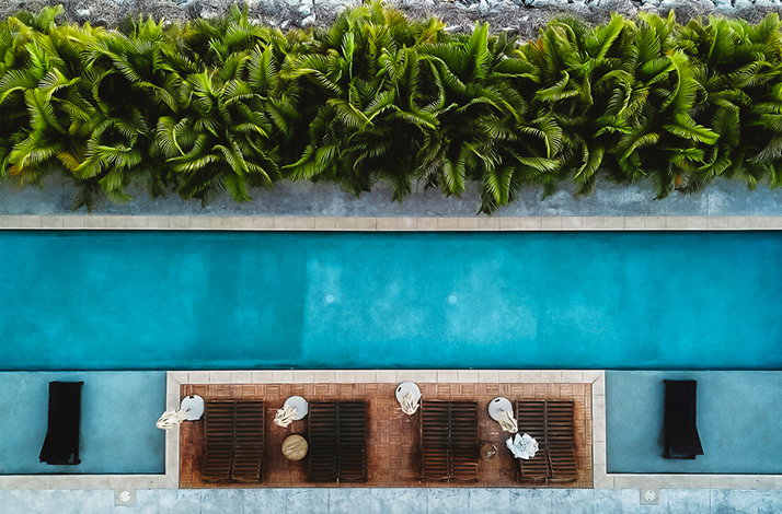 Overhead shot captures an outdoor pool area, featuring a long rectangular pool with lounge chairs and lush greenery