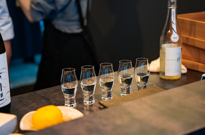 Five glasses of sake lined up on a counter with a sake bottle and a lemon.