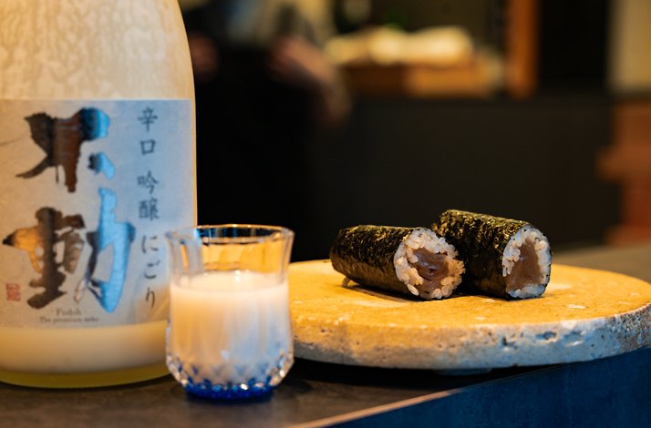 Two sushi rolls and a glass of cloudy sake on a round stone plate next to a sake bottle.